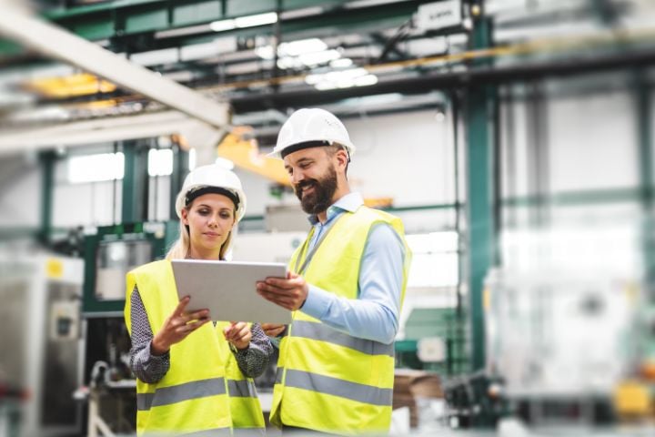 Two people standing in a production plant and holding a document in their hands.